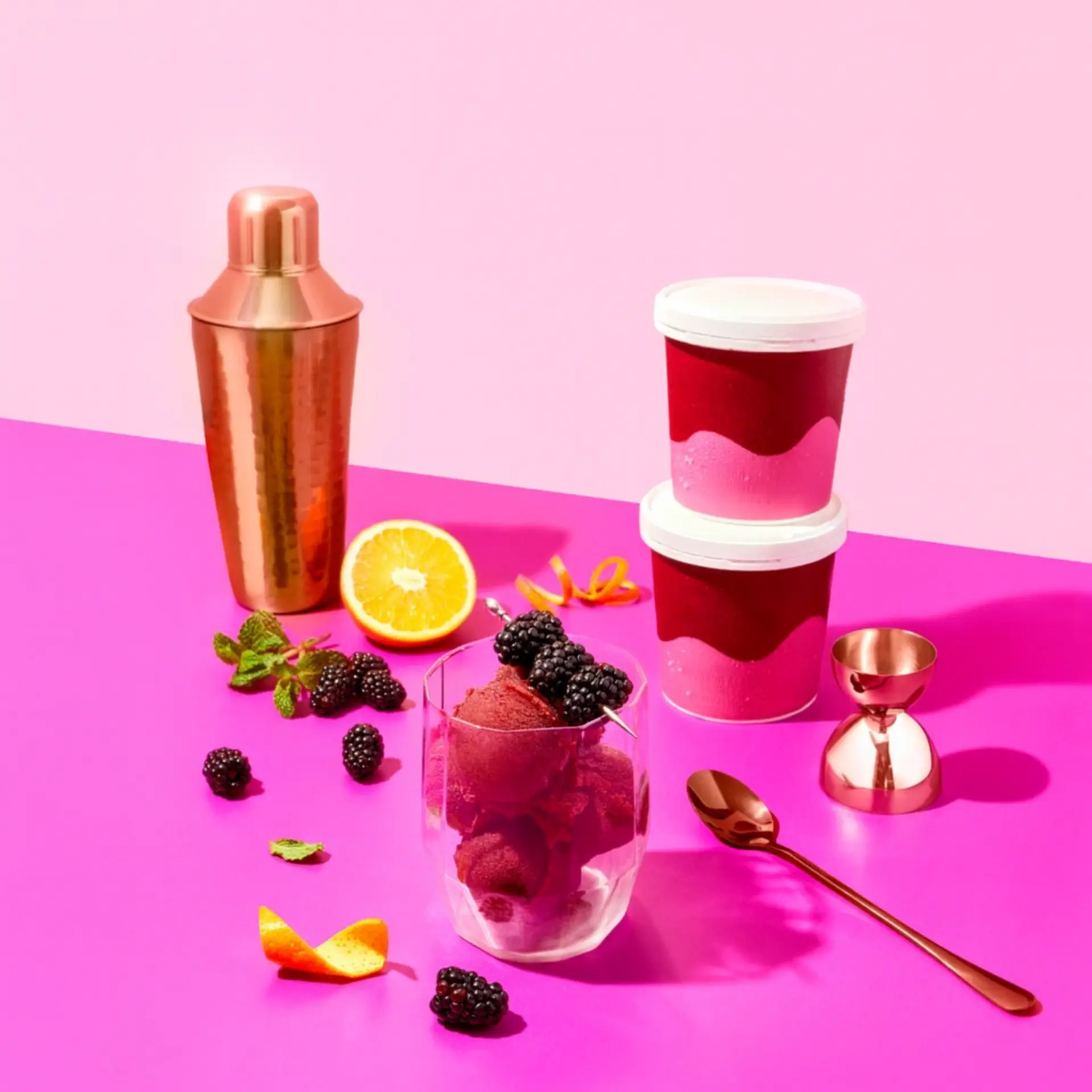 A clear glass with blackberry sorbet, blackberries, and orange peel sits on a pink surface, surrounded by two ice cream tubs, a copper cocktail shaker, jigger, spoon, mint, orange slice, and blackberries.