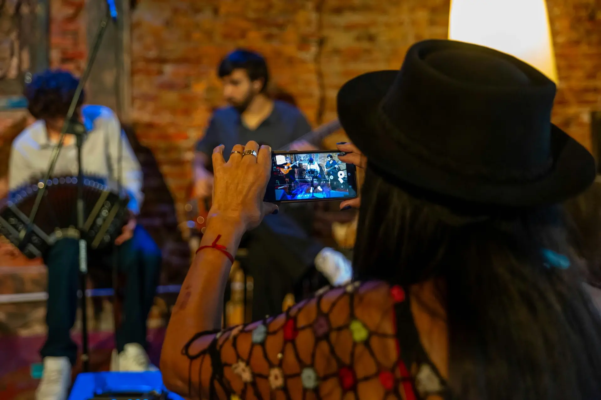 A person wearing a black hat takes a photo or video of two musicians seated in front of a brick wall using a smartphone. The musicians and background are out of focus, while the phone screen is clear.