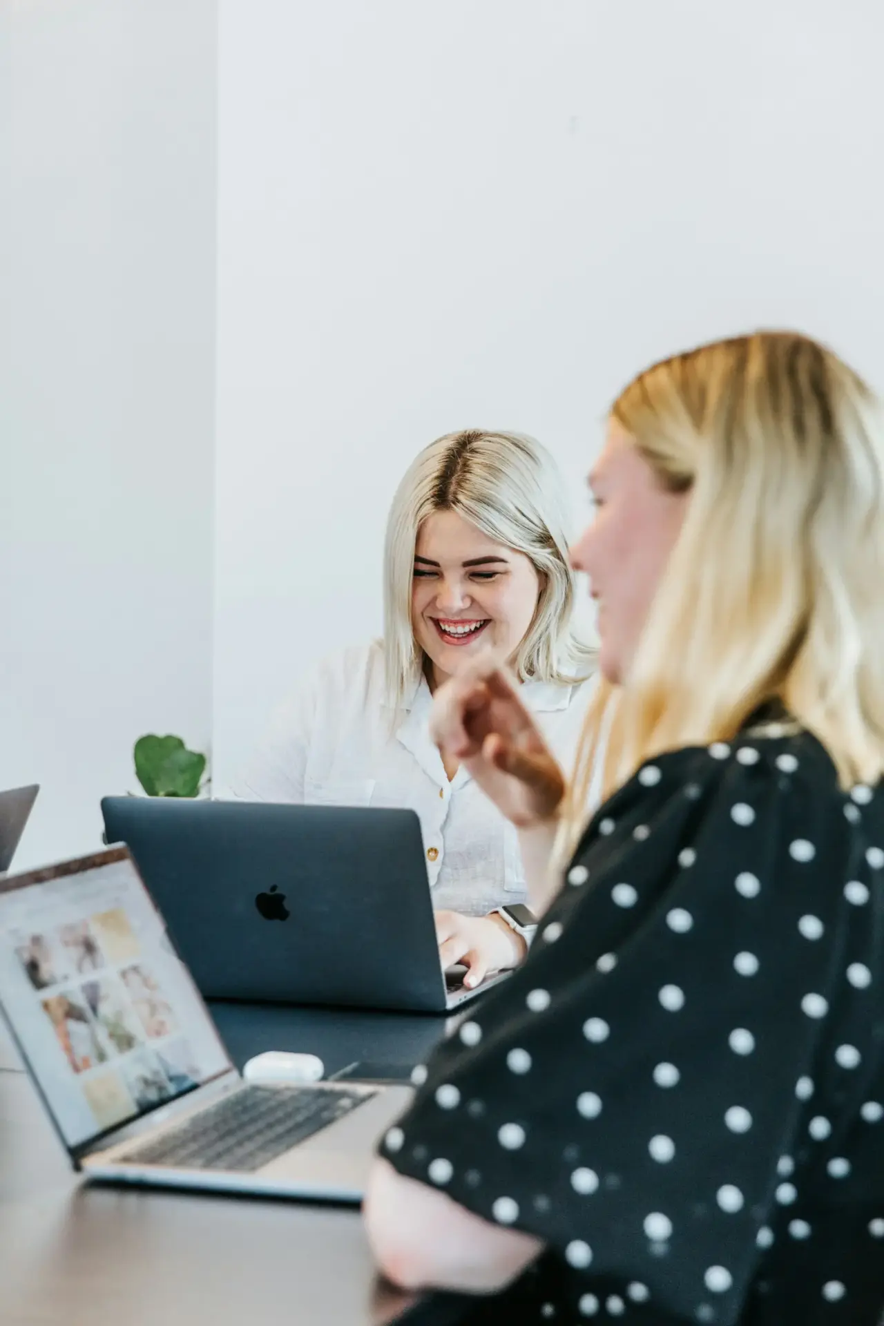 Two women with blonde hair are sitting at a table using laptops. One woman in the background is smiling, while the woman in the foreground is gesturing, with her face slightly out of focus.